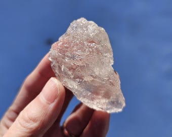 May include: A clear, translucent crystal with a rough, irregular surface. The crystal is held in a hand against a blue background.