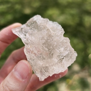 Può includere: Un grande cristallo trasparente e traslucido con una superficie ruvida e irregolare. Il cristallo è tenuto in una mano, e lo sfondo è sfocato.