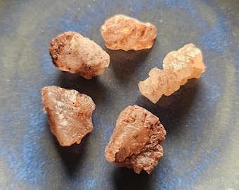 May include: Five rough-cut, translucent, light-orange crystals are arranged on a blue ceramic plate. The crystals have a slightly rough texture and varying shapes. The plate has a dark blue rim and a lighter blue center, creating a contrasting background.