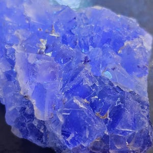 May include: Close-up of a vibrant blue fluorite mineral specimen. The crystalline structure is visible, with sharp, angular formations and a deep, saturated color. The surface reflects light, creating a shimmering effect.
