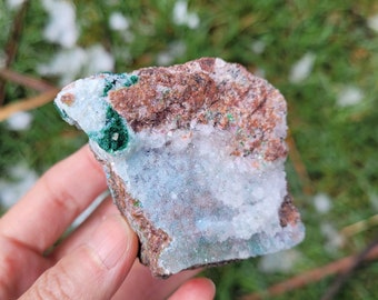 May include: A raw mineral specimen featuring a cluster of translucent white crystals embedded in a matrix of reddish-brown rock. The stone displays vibrant patches of deep green malachite and subtle blue mineral deposits across its textured surface.