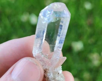 May include: A clear, elongated quartz crystal point with a hexagonal structure and a faceted termination. The crystal exhibits internal fractures and a subtle iridescent sheen, attached to a cluster of smaller, opaque white mineral base crystals.