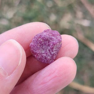 May include: A small, raw, deep magenta-colored gemstone with a rough, textured surface and irregular, rounded edges. The stone displays a vibrant, saturated pinkish-red hue throughout its crystalline structure, showing natural facets and mineral inclusions.