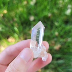 May include: A clear quartz crystal with a pointed tip and a rough base. The crystal is held against a blurred green background, showcasing its translucent quality and natural form. The crystal is approximately 2 inches in length.