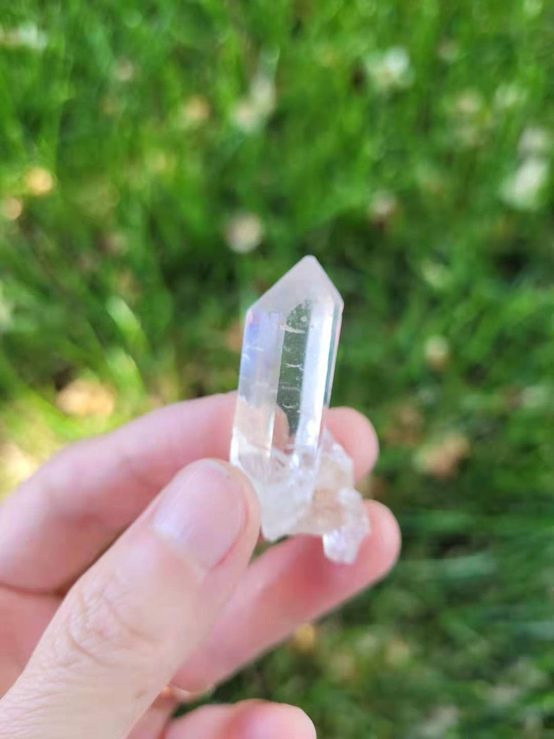 May include: A clear quartz crystal with a pointed tip and a rough base. The crystal is held against a blurred green background, showcasing its translucent quality and natural form. The crystal is approximately 2 inches in length.