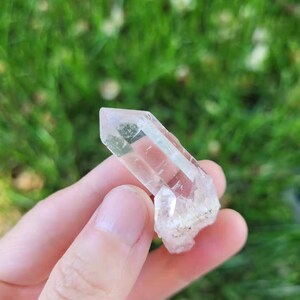 May include: A clear quartz crystal with a pointed tip and a rough, textured base. The crystal is held in a hand against a blurred green background of grass.