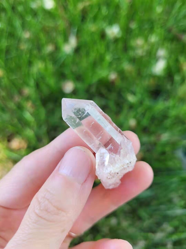May include: A clear quartz crystal with a pointed tip and a rough, textured base. The crystal is held in a hand against a blurred green background of grass.
