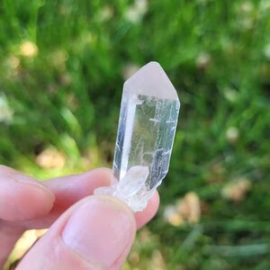 May include: A clear, elongated crystal with a pale pink tip. The crystal is held against a blurred green background, suggesting an outdoor setting. The crystal has a natural, rough texture.