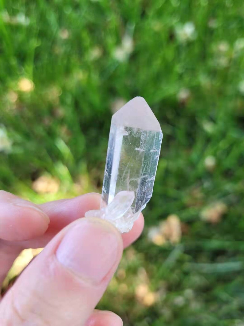 May include: A clear, elongated crystal with a pale pink tip. The crystal is held against a blurred green background, suggesting an outdoor setting. The crystal has a natural, rough texture.