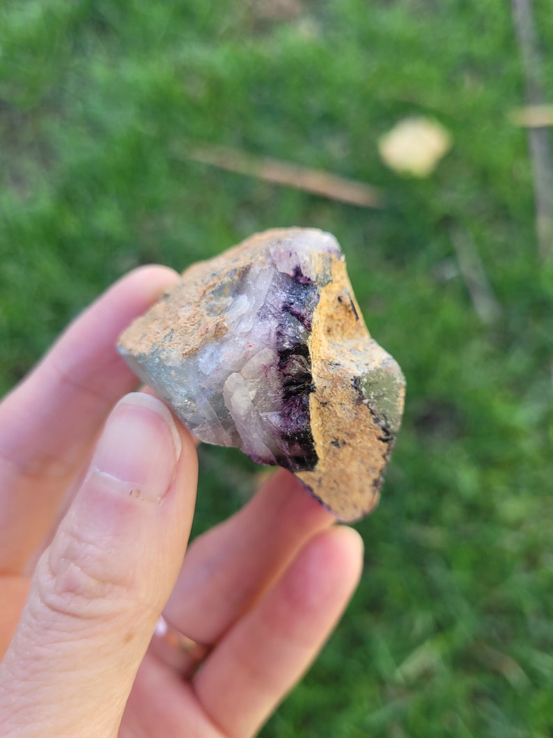 May include: A raw mineral specimen featuring a combination of translucent white crystalline structures and a deep purple band, encased within a rough, textured tan and brown rocky matrix with visible earthy deposits on the exterior surface.