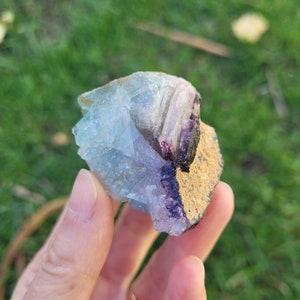 May include: A raw mineral specimen featuring translucent light blue and pale purple crystalline structures. One side of the stone displays a rough, textured, tan-colored matrix with a deep purple mineral deposit along the edge of the crystal formation.