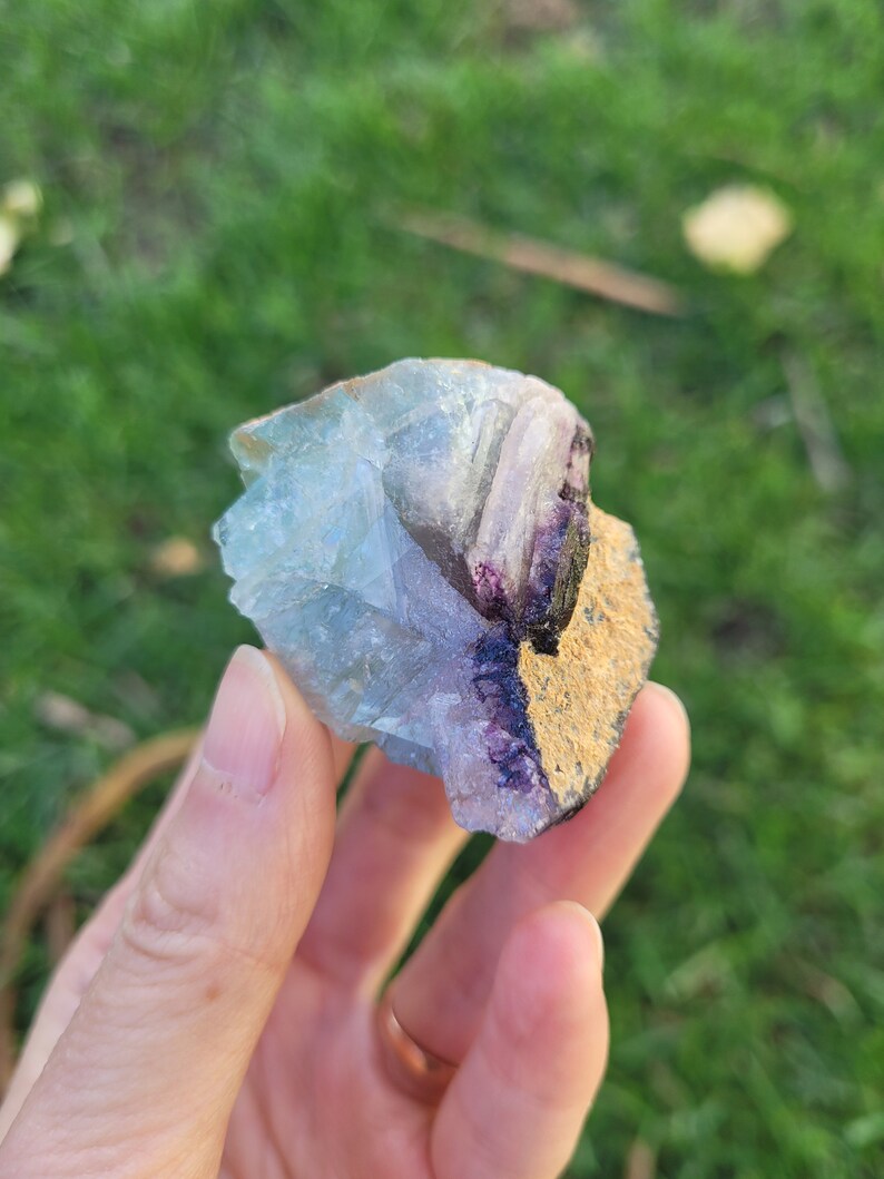 May include: A raw mineral specimen featuring translucent light blue and pale purple crystalline structures. One side of the stone displays a rough, textured, tan-colored matrix with a deep purple mineral deposit along the edge of the crystal formation.