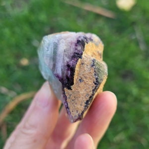 May include: A raw fluorite crystal specimen featuring a combination of translucent light blue and deep purple crystalline structures alongside a textured, tan-colored mineral matrix on one side, showcasing natural geometric facets and edges.