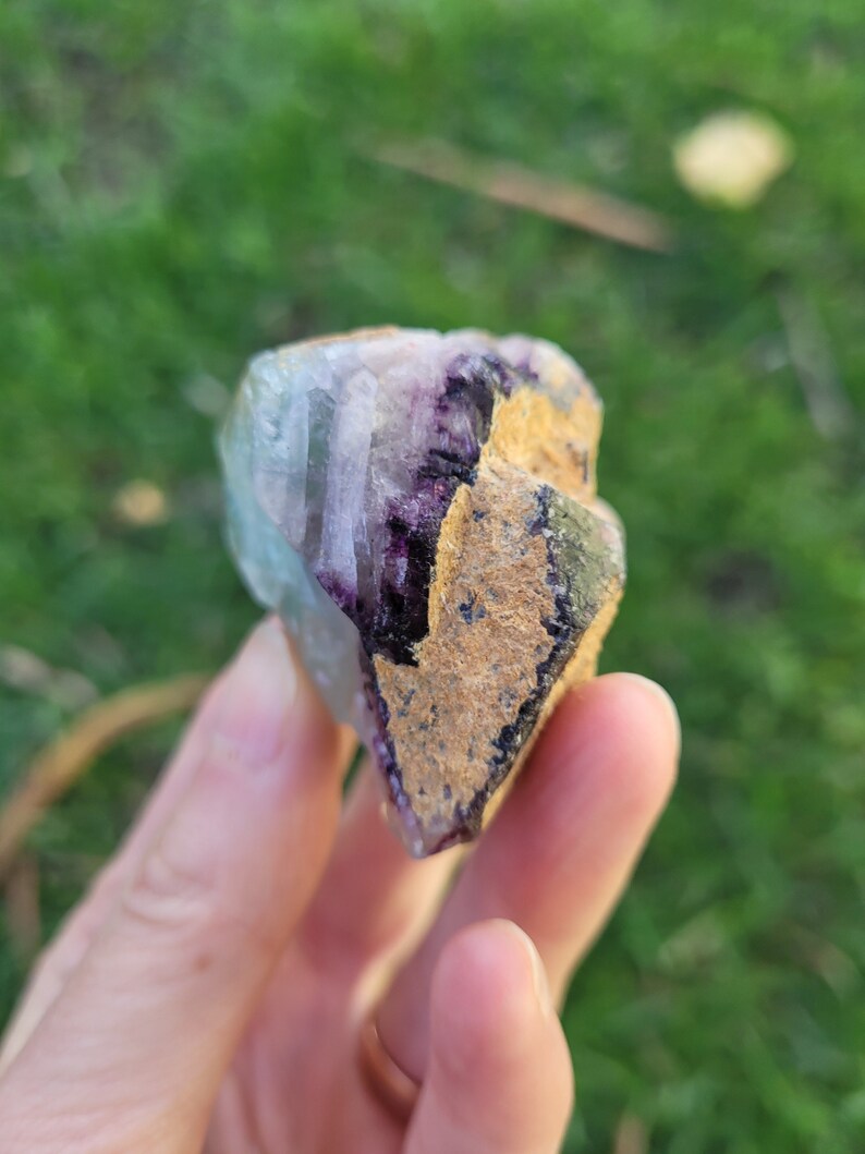 May include: A raw fluorite crystal specimen featuring a combination of translucent light blue and deep purple crystalline structures alongside a textured, tan-colored mineral matrix on one side, showcasing natural geometric facets and edges.