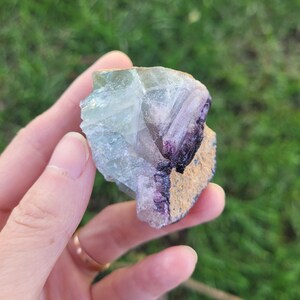 May include: A raw fluorite crystal specimen featuring translucent pale green and clear sections alongside a deep purple band and a textured, tan-colored mineral matrix edge. The stone displays natural geometric facets and crystalline structures.