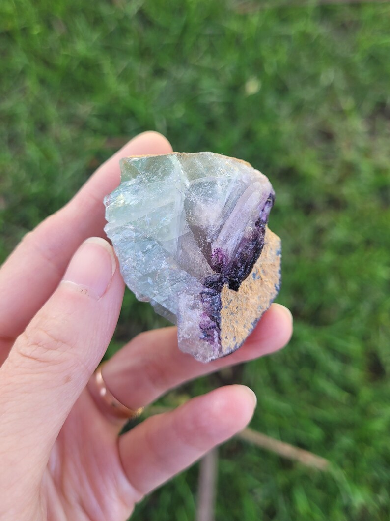 May include: A raw fluorite crystal specimen featuring translucent pale green and clear sections alongside a deep purple band and a textured, tan-colored mineral matrix edge. The stone displays natural geometric facets and crystalline structures.