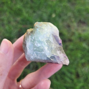 May include: A raw, translucent fluorite crystal specimen featuring a mix of pale green, light blue, and subtle purple hues. The mineral has a jagged, multi-faceted surface with visible internal fractures and a glassy, reflective texture throughout.