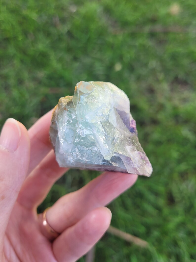 May include: A raw, translucent fluorite crystal specimen featuring a mix of pale green, light blue, and subtle purple hues. The mineral has a jagged, multi-faceted surface with visible internal fractures and a glassy, reflective texture throughout.
