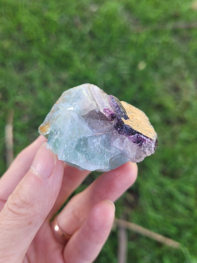May include: A raw fluorite crystal specimen featuring translucent light green and blue hues with a distinct patch of deep purple coloration and a textured, tan-colored mineral matrix on one side, showcasing natural geometric crystalline facets.
