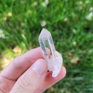 May include: A clear quartz crystal with a pointed tip and a rough, textured base. The crystal is held against a backdrop of green grass, showcasing its natural clarity and form. This crystal is a natural gemstone.