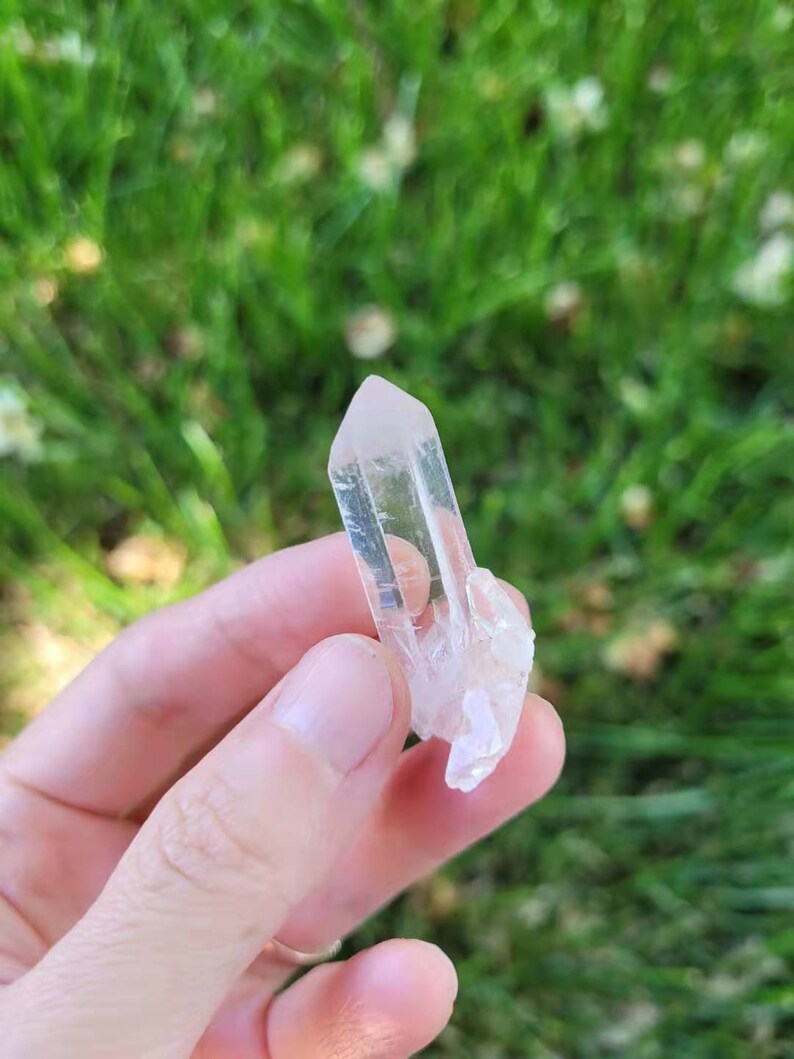 May include: A clear quartz crystal with a pointed tip and a rough, textured base. The crystal is held against a backdrop of green grass, showcasing its natural clarity and form. This crystal is a natural gemstone.