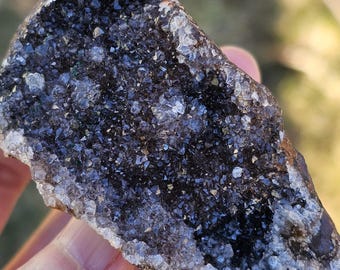 Op de afbeelding: Een close-up van een donkergrijze en zwarte kristal met een witte, glinsterende rand. De kristal is ruw en heeft een natuurlijke, onbewerkte uitstraling.