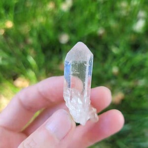 May include: A clear quartz crystal with a pointed tip and a rough base. The crystal is held in a hand against a blurred green background. The quartz is translucent, allowing light to pass through.