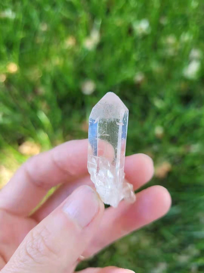 May include: A clear quartz crystal with a pointed tip and a rough base. The crystal is held in a hand against a blurred green background. The quartz is translucent, allowing light to pass through.