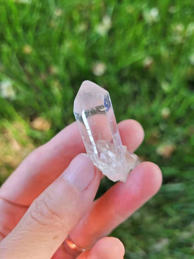 May include: A clear quartz crystal point with a natural, rough base. The crystal is held in a hand against a blurred green background. The quartz is transparent with a slight pinkish hue.