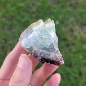 May include: A raw, translucent mineral specimen featuring a gradient of colors, transitioning from a clear, pale mint green at the top to a deep, saturated purple at the base, with visible internal fractures and a jagged, crystalline texture.