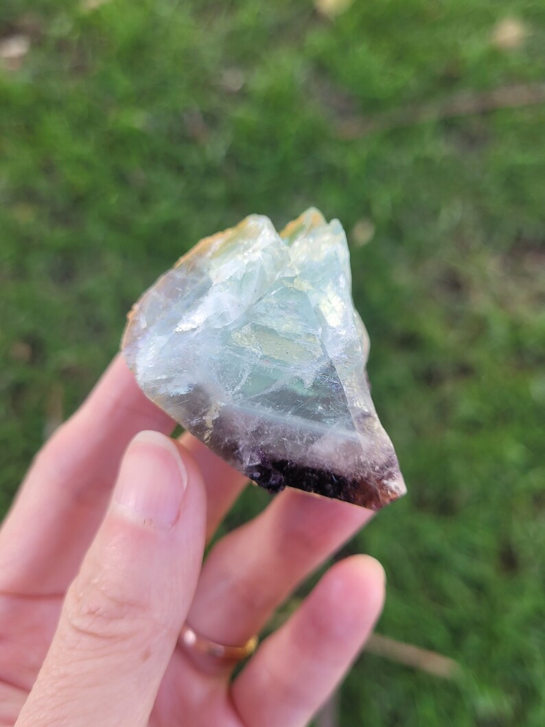 May include: A raw, translucent mineral specimen featuring a gradient of colors, transitioning from a clear, pale mint green at the top to a deep, saturated purple at the base, with visible internal fractures and a jagged, crystalline texture.