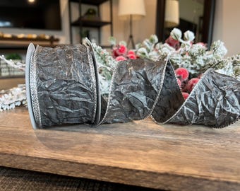 Puede incluir: Un rollo de cinta gris oscuro texturizada con un borde plateado. La cinta está parcialmente desenrollada, revelando su superficie arrugada. Ideal para envolver regalos o decoraciones festivas.