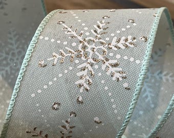 Puede incluir: Una cinta azul claro con un diseño de copo de nieve blanco y detalles de purpurina plateada. La cinta tiene un borde con alambre.