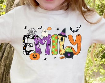 Może przedstawiać: Kremowy t-shirt z napisem "Emily" w halloweenowym wzorze. Litery ozdobione są nietoperzami, kapeluszem czarownicy, duchem, dynią i kociołkiem. Koszulka jest noszona przez dziecko.