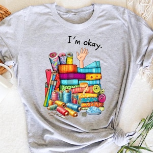 Peut inclure: T-shirt gris avec une illustration colorée de chutes de tissu, de bobines de fil, de ciseaux et une main qui sort de la pile. Le texte "I'm okay." est imprimé au-dessus de l'illustration.