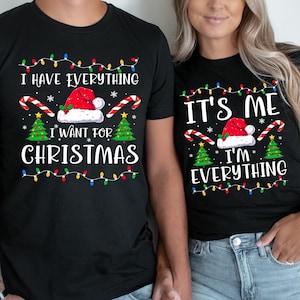 May include: Black Christmas-themed t-shirts with matching designs. One shirt reads "I HAVE EVERYTHING I WANT FOR CHRISTMAS" and the other says "IT'S ME I'M EVERYTHING". Both shirts feature a Santa hat, candy canes, Christmas trees, and string lights.