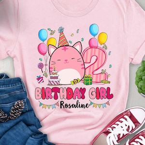 Puede incluir: Camiseta rosa con un gato de dibujos animados que lleva un gorro de fiesta y sostiene un pastel. La camiseta dice "Birthday Girl" y tiene un número 2. El nombre "Rosaline" está escrito debajo del texto.