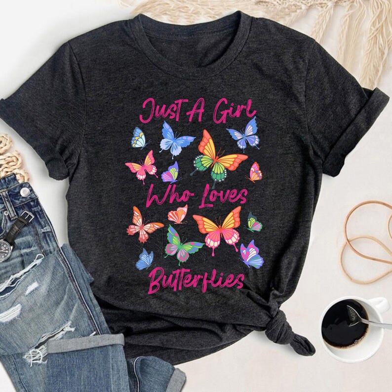 May include: A dark gray t-shirt with the text "Just A Girl Who Loves Butterflies" printed in pink. The shirt features a colorful butterfly design.