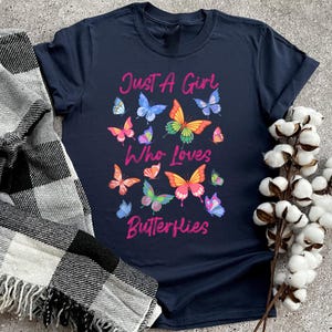 May include: A navy blue t-shirt with the text "Just a Girl Who Loves Butterflies" in pink lettering. The shirt is decorated with colorful butterfly graphics.