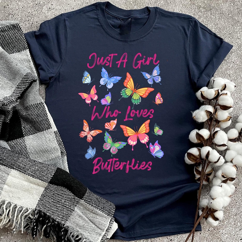 May include: A navy blue t-shirt with the text "Just a Girl Who Loves Butterflies" in pink lettering. The shirt is decorated with colorful butterfly graphics.