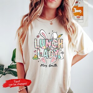 May include: A light beige t-shirt with a colorful graphic of bunny ears, the words "Lunch Lady", and a paw print. The text "Mrs. Smith" is below the graphic.
