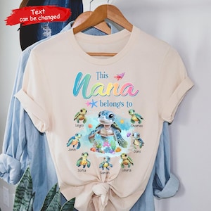 以下が含まれることがあります： 薄いベージュのTシャツに「This Nana belongs to」という文字と、それぞれの名前が書かれたカラフルなウミガメのグラフィックが描かれています。名前は、レイラ、ジョン、ソフィア、サラ、ローラ、チャールズ、ダニです。
