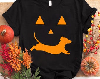 May include: Black t-shirt featuring a Halloween-themed design. The design includes an orange pumpkin face and a running dachshund dog silhouette. The shirt is surrounded by fall foliage and pumpkins.
