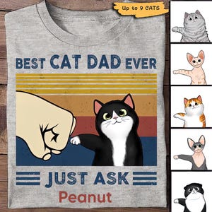Può includere: Una maglietta grigia con una grafica in stile vintage che dice "Best Cat Dad Ever" e "Just Ask Peanut". La grafica presenta un gatto bianco e nero con un pugno chiuso.
