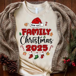 May include: A cream-coloured t-shirt with the text "FAMILY Christmas 2025" in red and black. The design includes a Santa hat, Christmas stockings, candy canes, and a gingerbread man. The shirt is displayed on a wooden surface.