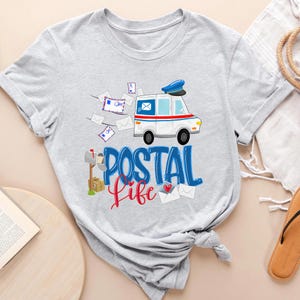 May include: A gray t-shirt with a white mail truck, a mailbox, and the text "Postal Life" in blue and red letters. The mail truck has a blue hat on top and is surrounded by envelopes.