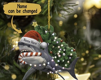 Puede incluir: Adorno navideño de tiburón con gorro de Papá Noel y bastón de caramelo. El tiburón es gris y blanco, con el texto "Name Here" en el gorro. El adorno incluye un pequeño árbol de Navidad y luces de cadena. También está presente el texto "Name can be changed".