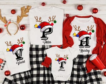 Pode incluir: Um conjunto de camisetas e pijamas de Natal brancos com um design de rena e chapéu de Papai Noel. Cada camisa apresenta uma letra e um nome: Violet, Peter, Harley e Thomas. Os pijamas são xadrez preto e branco.