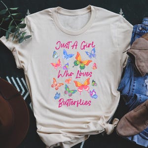 May include: A beige t-shirt with the text "Just a Girl Who Loves Butterflies" printed in pink. The shirt is decorated with colorful butterfly illustrations.