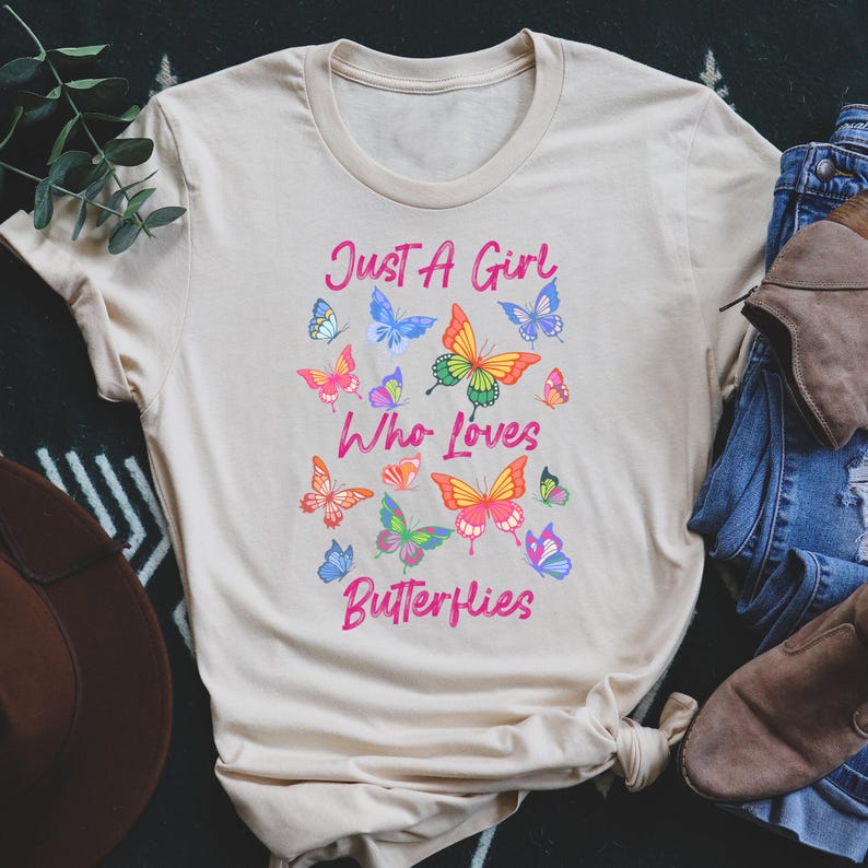 May include: A beige t-shirt with the text "Just a Girl Who Loves Butterflies" printed in pink. The shirt is decorated with colorful butterfly illustrations.